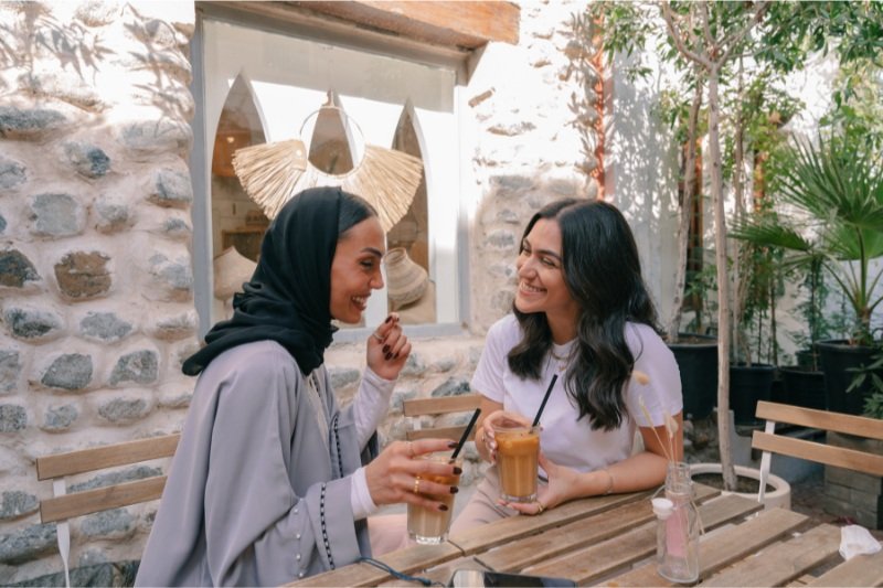 Two women drinking coffee and sitting at an outdoor table while smiling.