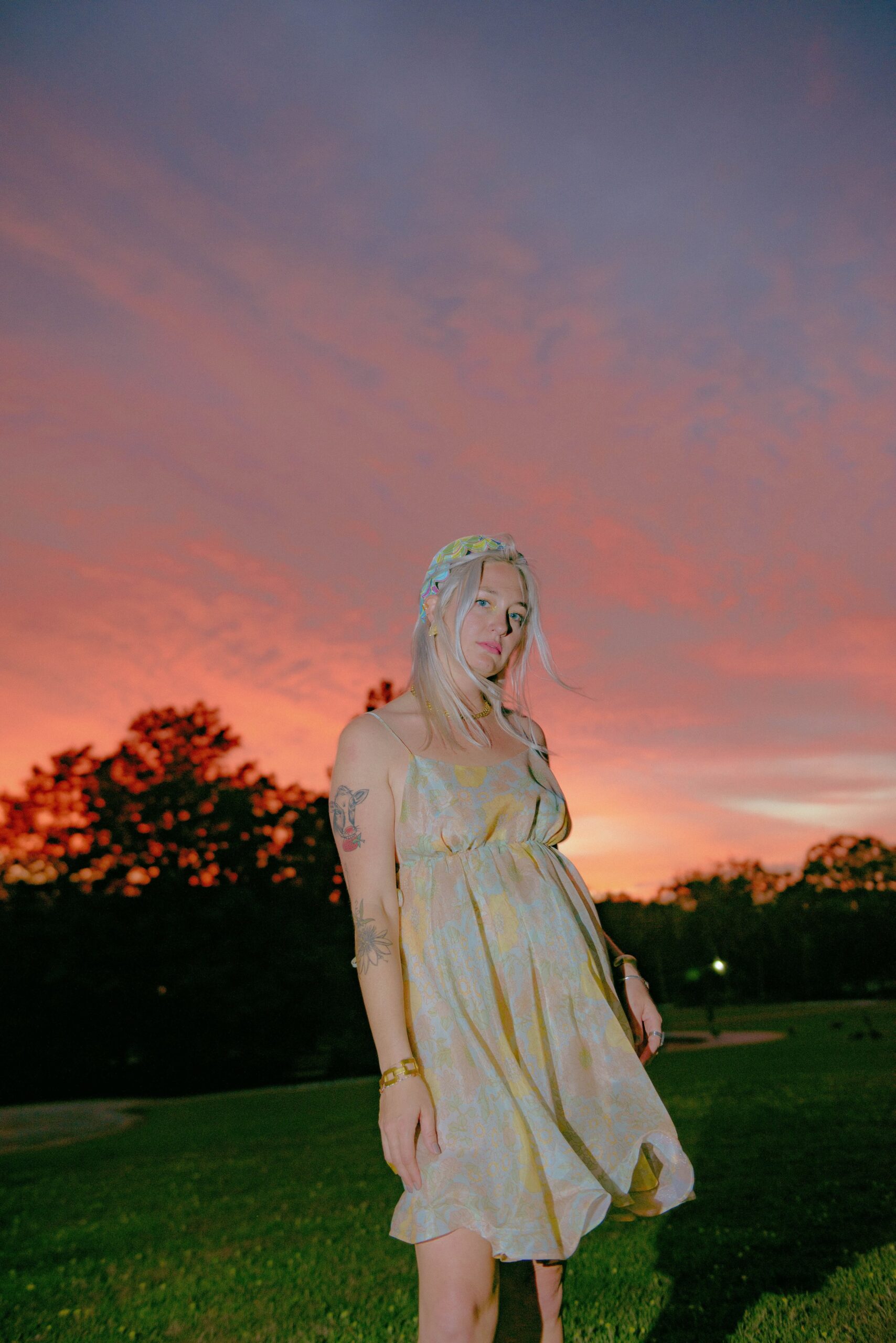 Woman wearing a sundress and flower crown stands in a field at sunset.